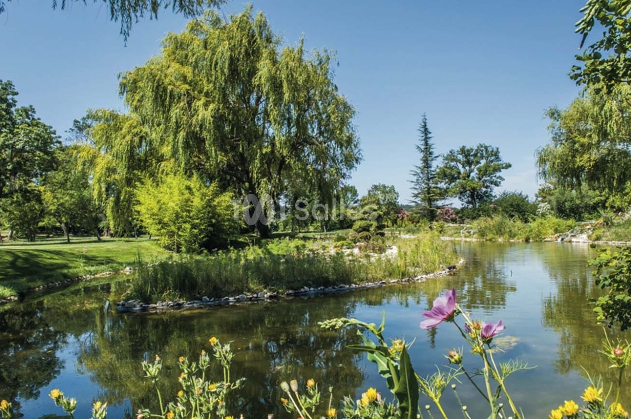 Étang entouré de verdure avec des arbres, des fleurs au premier plan et un ciel bleu dégagé.