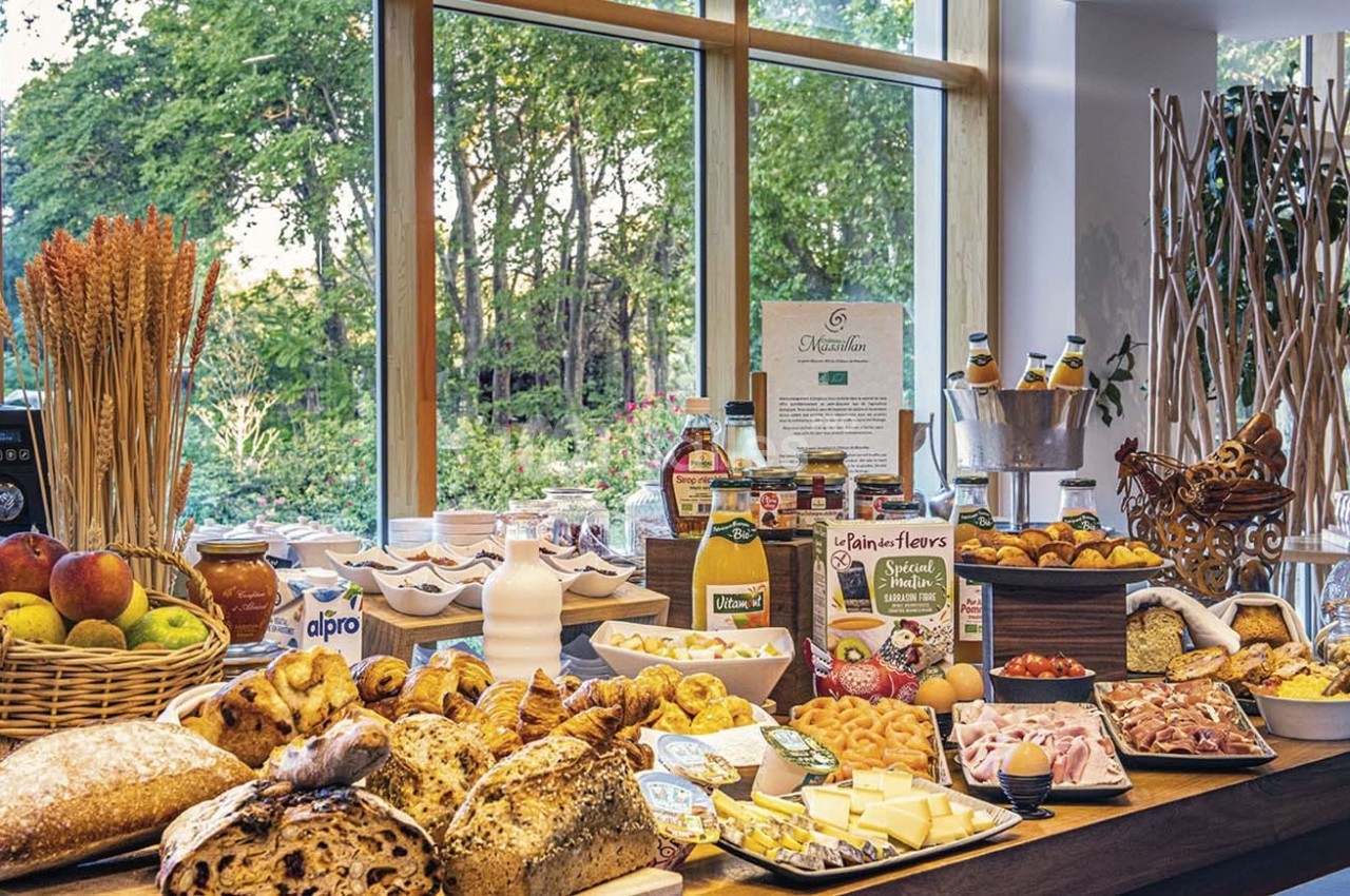 Buffet de petit-déjeuner avec pains, viennoiseries, fruits, charcuteries, fromages, jus et céréales, devant une baie vitrée.