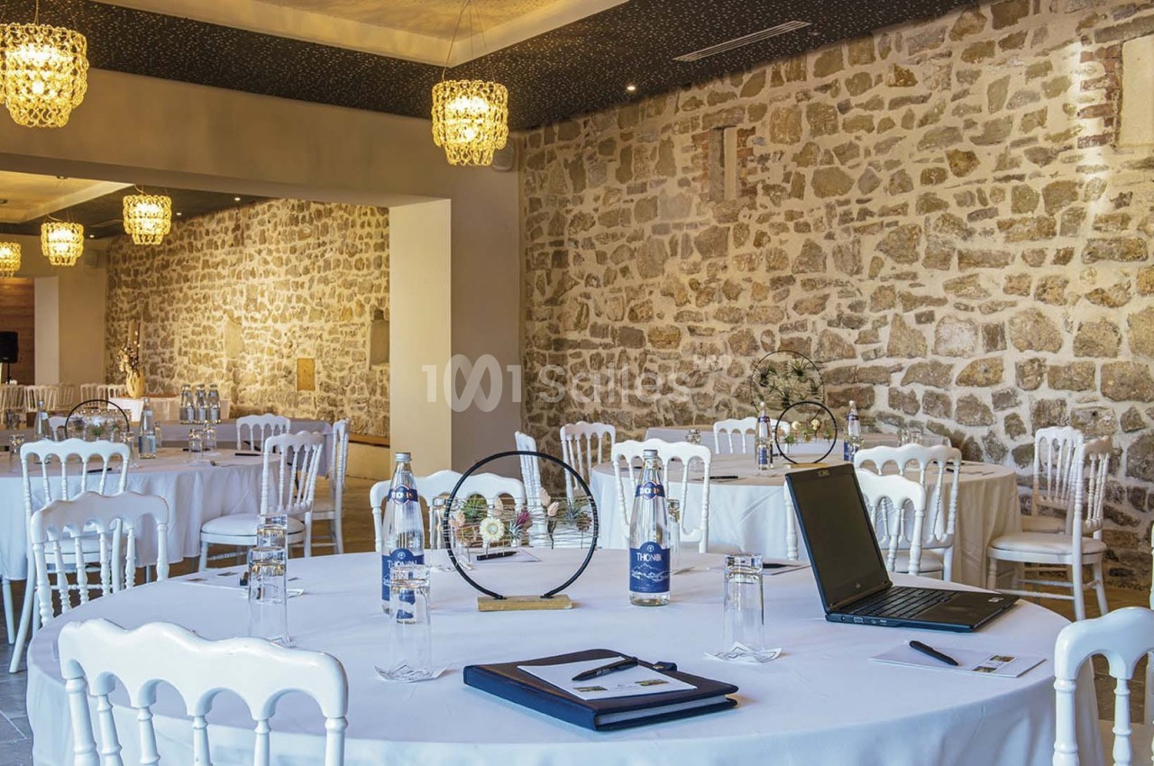 Salle de réception avec murs en pierre, tables rondes dressées et décorations lumineuses suspendues.