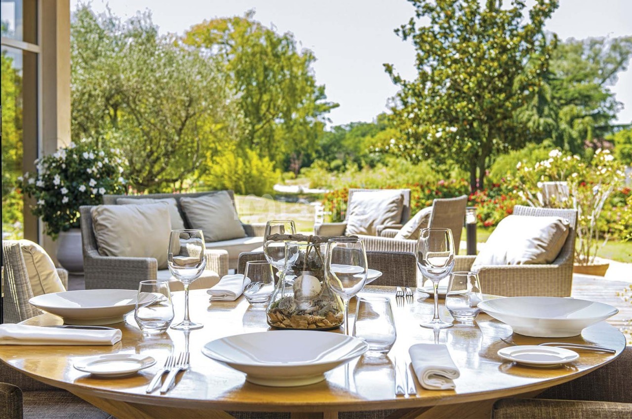 Table dressée avec vaisselle blanche et verres, donnant sur un jardin verdoyant avec mobilier d'extérieur.