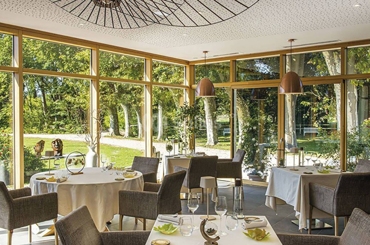 Salle de restaurant lumineuse avec de grandes baies vitrées donnant sur un jardin arboré.