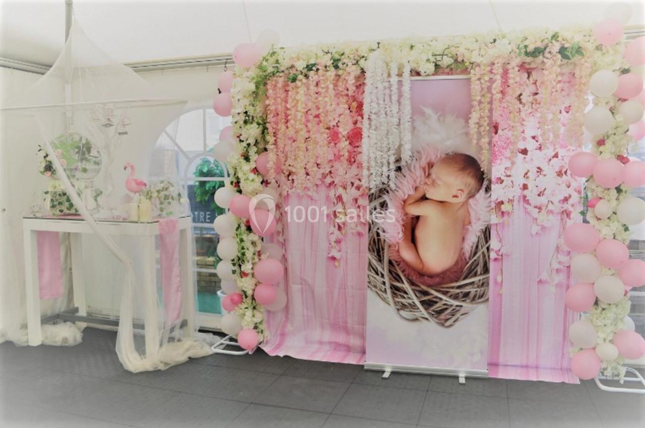 Décoration de fête avec un panneau montrant un bébé dans un nid, entouré de fleurs, ballons roses et voilage blanc.