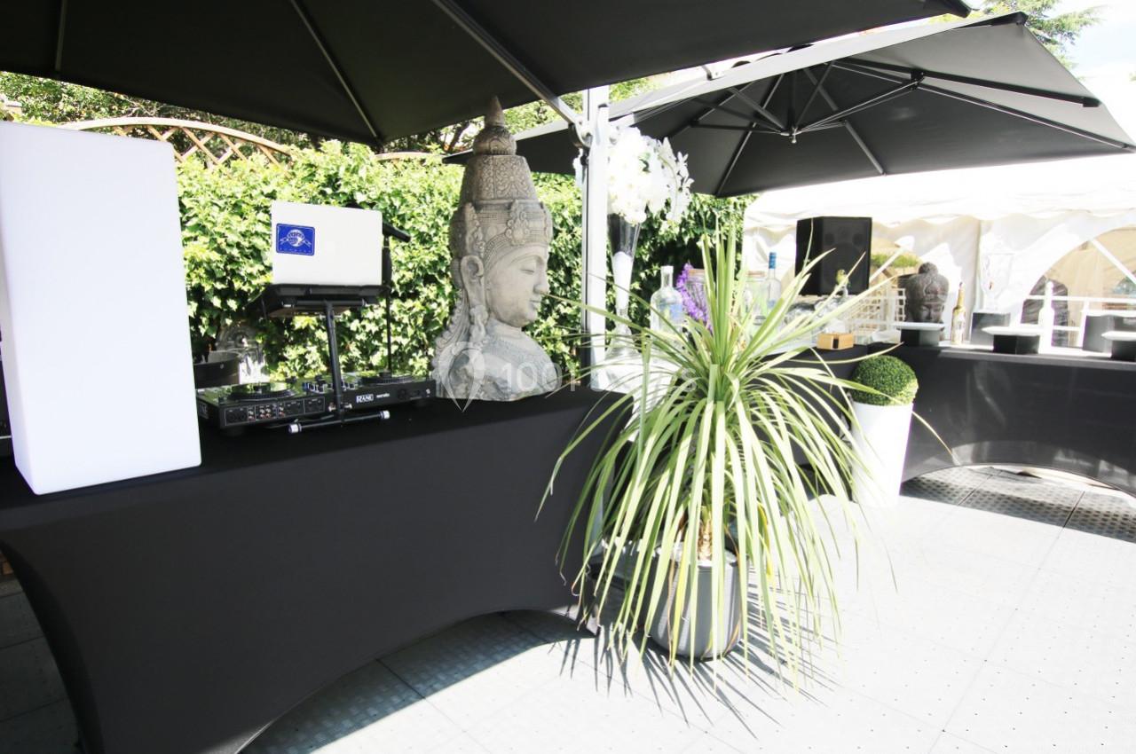 Espace extérieur avec table équipée de matériel audio, statue de Bouddha, plantes et parasols noirs.