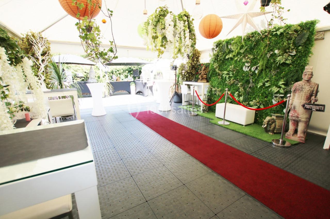 Espace événementiel décoré avec plantes, lanternes suspendues, tapis rouge et statue près d'une barrière.