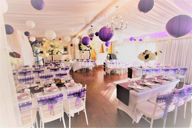 Salle de réception décorée avec des lanternes violettes, des fleurs blanches et des tables dressées pour un événement.