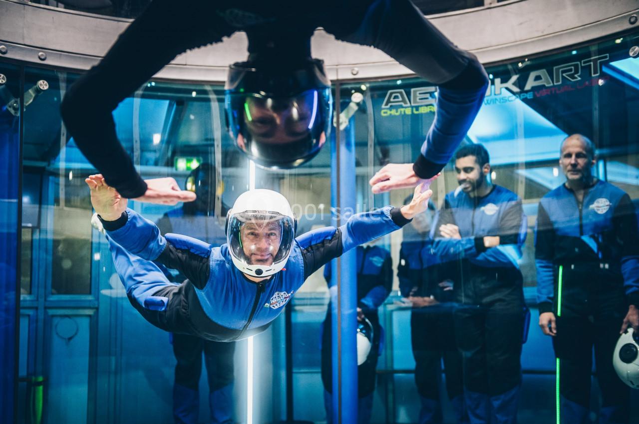 Un homme en combinaison et casque pratique la chute libre dans un simulateur, observé par des spectateurs en arrière-plan.
