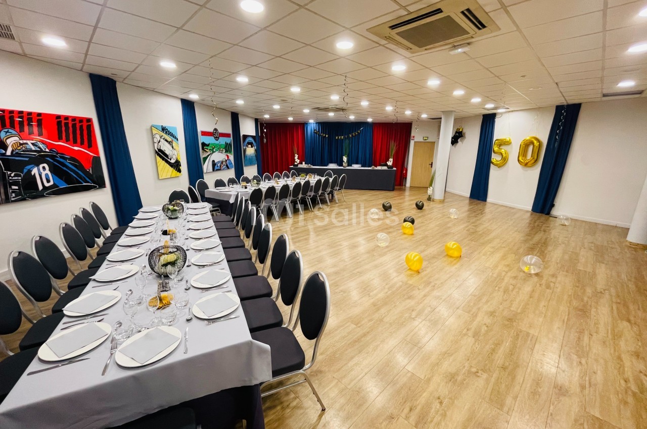Salle décorée pour un événement avec tables dressées, ballons dorés ’50’ et rideaux rouges en arrière-plan.