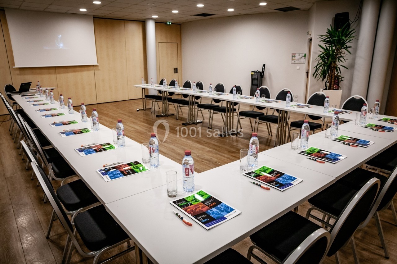 Salle de réunion avec tables en U, chaises, bouteilles d'eau, brochures colorées et écran de projection allumé.