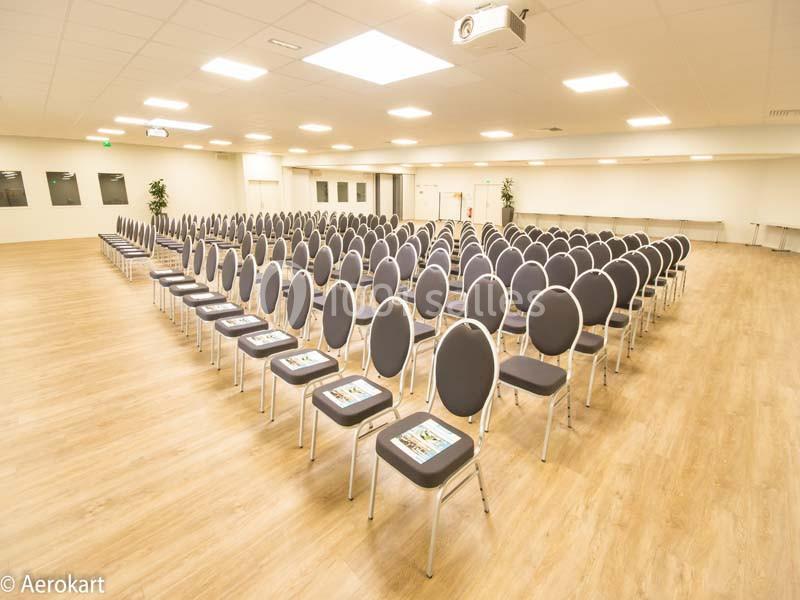 Salle de conférence lumineuse avec des rangées de chaises grises alignées sur un sol en parquet clair.