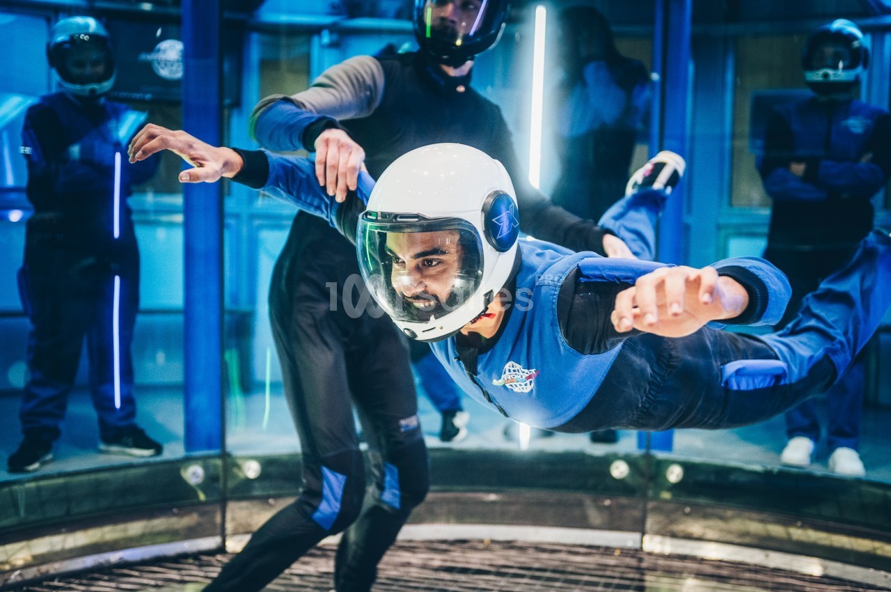 Un homme en combinaison et casque flotte dans un simulateur de chute libre, encadré par un instructeur.