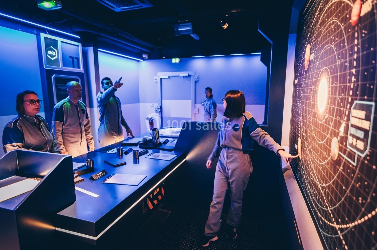 Des personnes en combinaison discutent et interagissent dans une salle futuriste équipée d'écrans et de consoles.