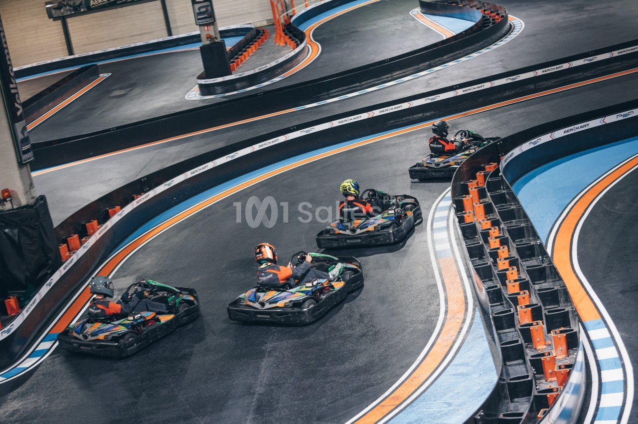 Quatre personnes conduisent des karts sur une piste intérieure avec des virages marqués et des bordures colorées.