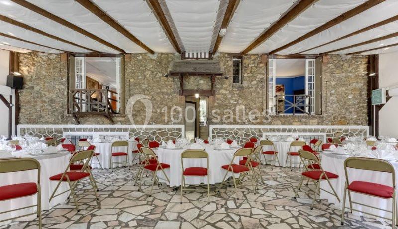 Salle de réception avec tables rondes dressées, chaises rouges et murs en pierre, sous un plafond à poutres apparentes.