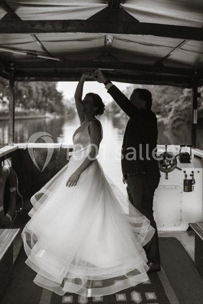 Un couple danse sur un bateau, la femme porte une robe de mariée fluide, l'homme un costume sombre.