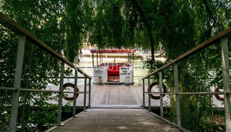 Passerelle en bois menant à un bateau amarré, entourée de végétation dense et ombragée.