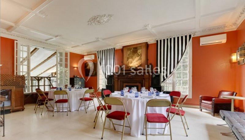 Salle lumineuse avec murs orange, tables rondes dressées, chaises pliantes rouges et cheminée ornée d'un tableau.