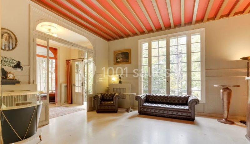 Salon lumineux avec canapés en cuir, grandes fenêtres, plafond à poutres rouges et décoration classique.