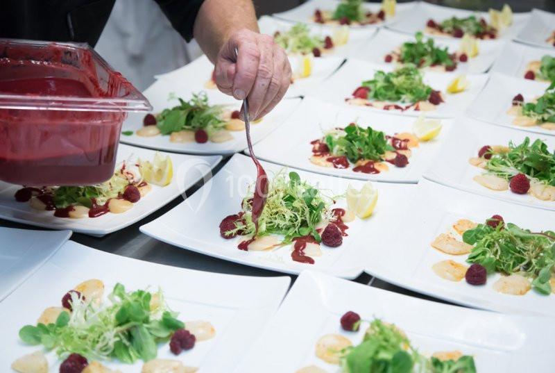 Un chef ajoute une sauce rouge sur des assiettes garnies de salade, framboises et tranches de fromage.
