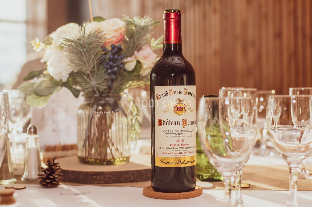 Bouteille de vin rouge posée sur une table dressée avec des verres, un vase de fleurs et une lumière naturelle.