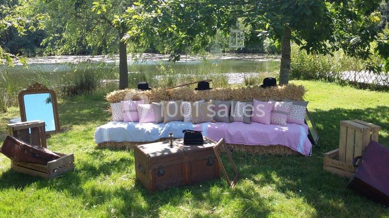 Banc de bottes de paille décoré de coussins et couvertures, entouré de valises et caisses en bois, près d'un étang.
