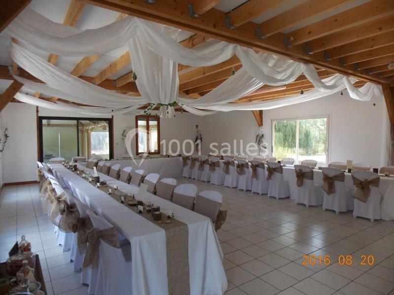 Salle de réception décorée avec des tables alignées, nappes blanches et voilages suspendus au plafond.