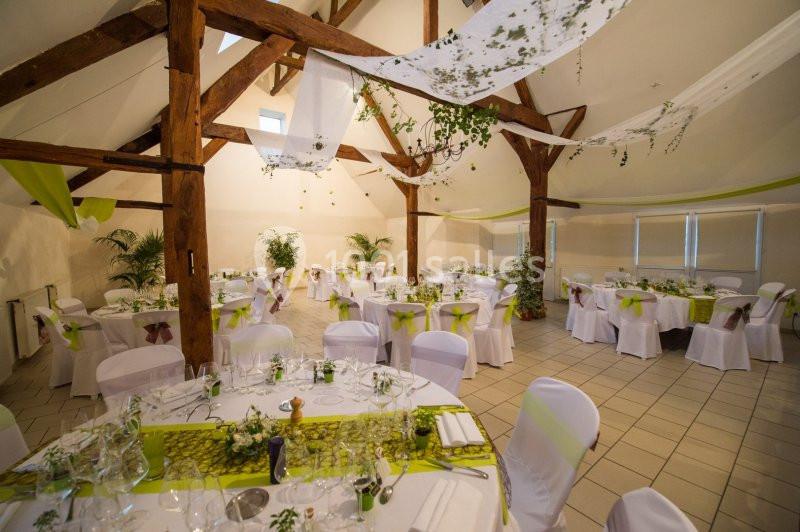 Salle de réception décorée pour un événement, avec tables rondes dressées, nappes blanches et ornements verts.