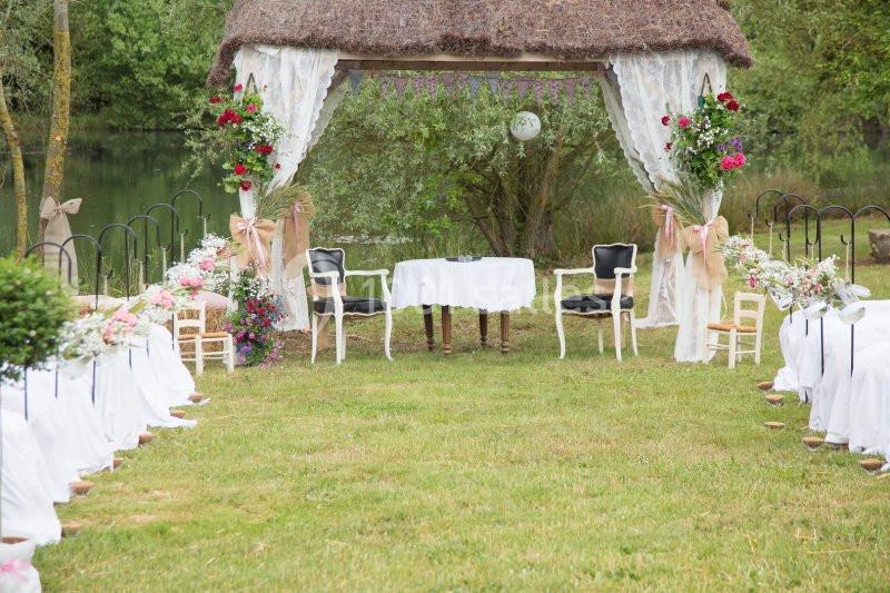Autel de mariage en plein air avec chaises décorées, arche fleurie et table blanche, entouré de verdure.