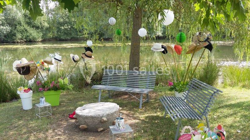 Deux bancs en métal sous un arbre près d'un étang, décorés de chapeaux, lanternes et fleurs.