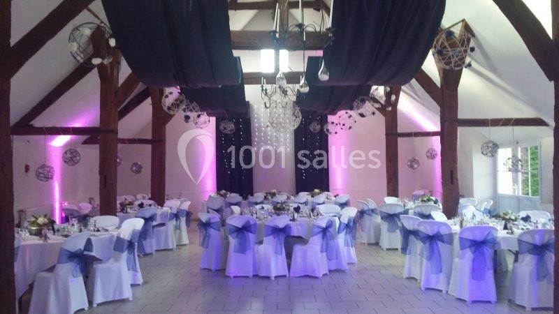 Salle de réception décorée avec des tables rondes, nappes blanches, chaises ornées de nœuds violets et éclairage tamisé.