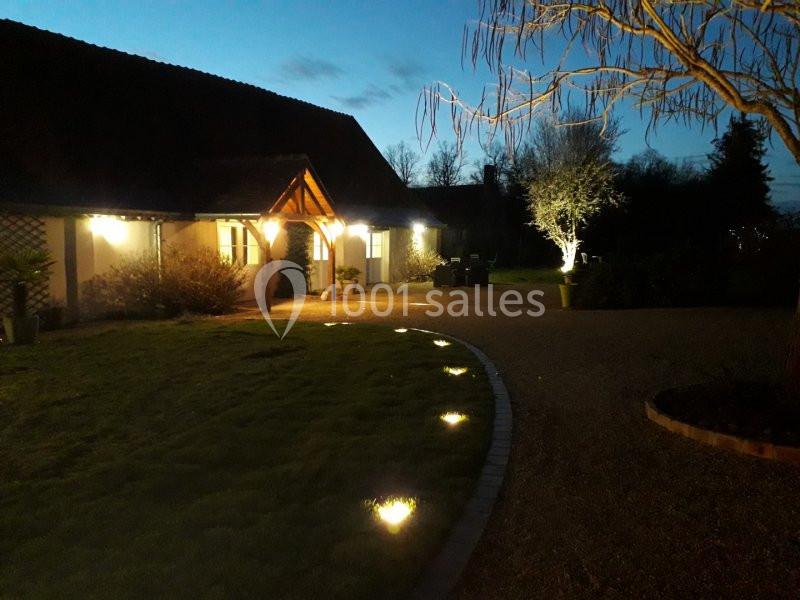 Maison éclairée à la tombée de la nuit, avec un chemin illuminé et un jardin arboré au premier plan.