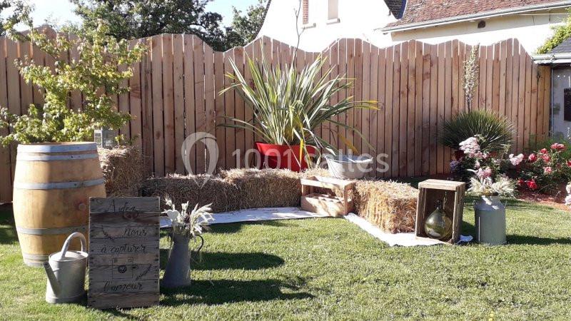Décor champêtre avec bottes de paille, tonneau en bois, plantes, arrosoirs et panneaux en bois sur une pelouse.