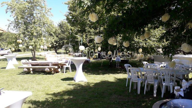 Jardin aménagé pour un événement avec tables, chaises blanches et décorations suspendues sous les arbres.