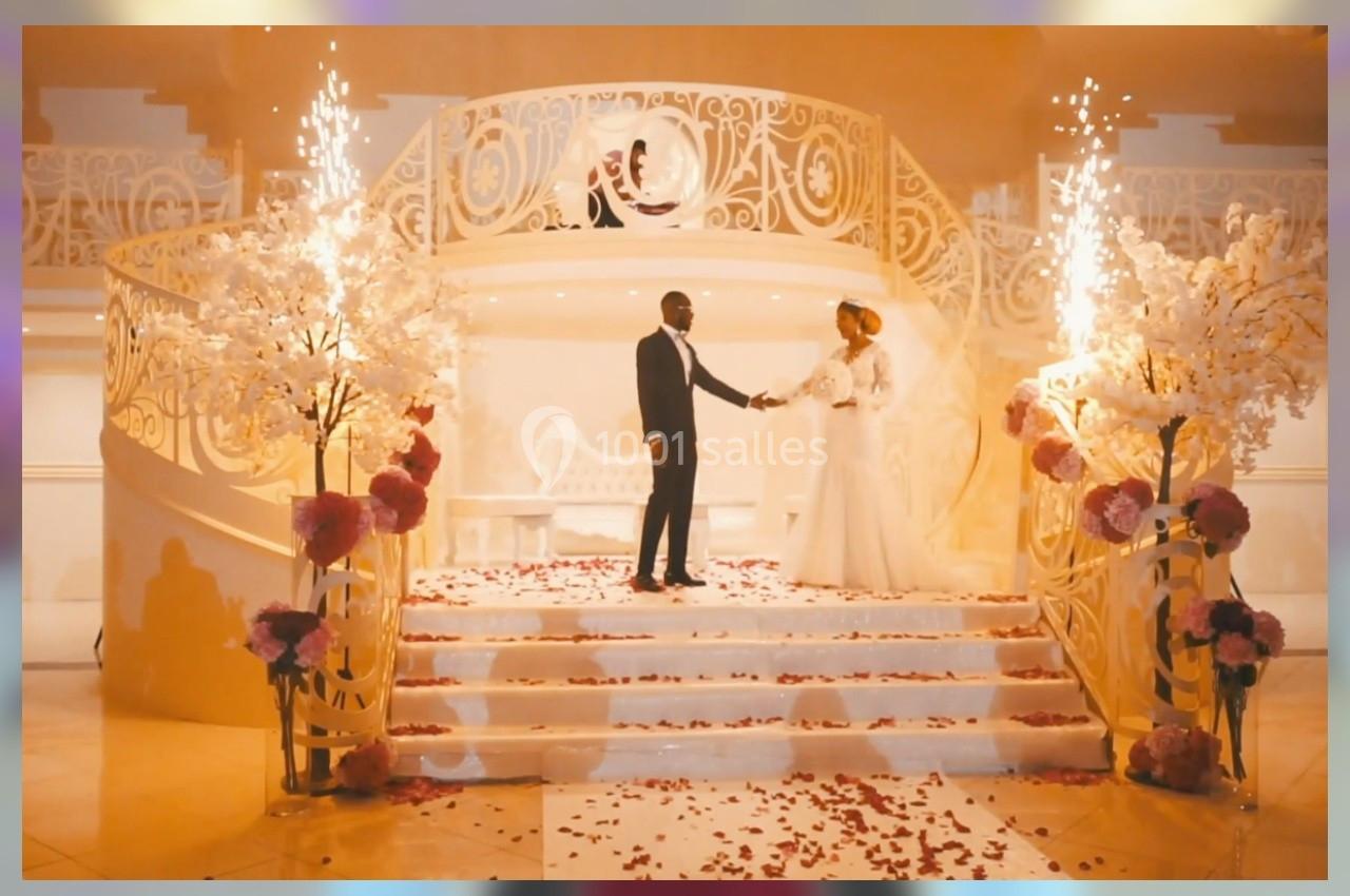 Un couple en tenue de mariage se tient sur un escalier décoré de fleurs et de lumières, entouré de jets d'étincelles.