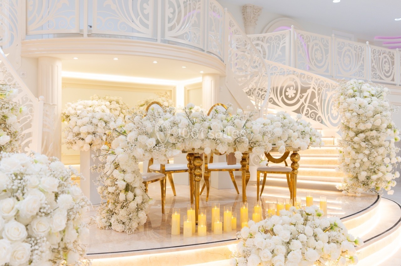 Salle de réception décorée de fleurs blanches, avec une table dorée et des bougies alignées sur des marches illuminées.