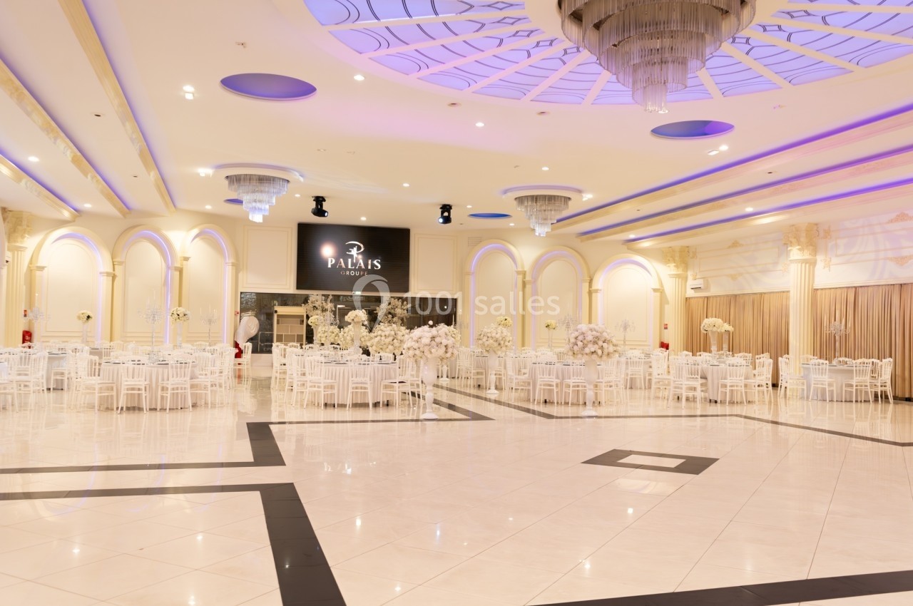 Salle de réception spacieuse avec tables rondes dressées, décorations florales blanches et éclairage élégant.