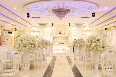 Salle de réception élégante avec tables décorées de nappes blanches, centres de table floraux et éclairage rose.
