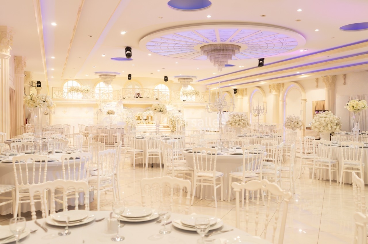 Salle de réception élégante avec tables rondes dressées, chaises blanches et décorations florales blanches.
