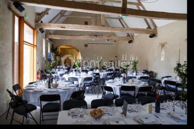 Salle en pierre avec poutres apparentes, tables rondes dressées et décorations lumineuses pour un événement.