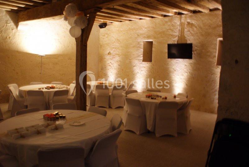 Salle en pierre avec poutres apparentes, tables rondes dressées et décorations lumineuses pour un événement.