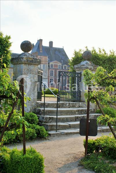 Entrée d'un jardin avec portail en fer forgé, escaliers en pierre et manoir en arrière-plan entouré de verdure.