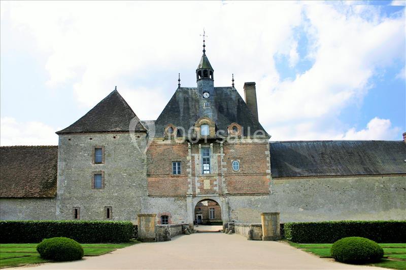 Façade d'un bâtiment historique en pierre avec une tour centrale, entouré de haies et d'un ciel partiellement nuageux.