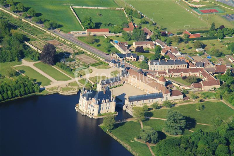 Vue aérienne d'un château entouré d'un lac, de jardins aménagés et d'un village avec des terrains verts.