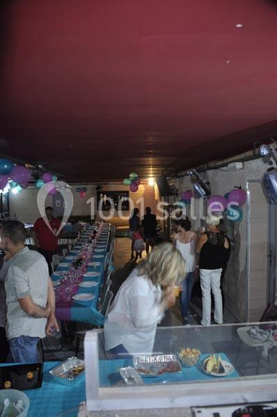 Salle décorée avec des ballons, tables dressées pour un repas, quelques personnes discutant et se servant au buffet.