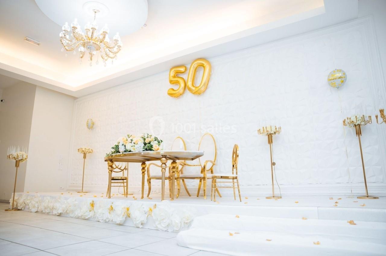 Location salle Viry-Châtillon (Essonne) - Glodie Evènements #4 Salle décorée en blanc et or avec une table, des chaises dorées, des ballons ’50’ et des chandeliers élégants.