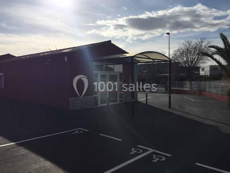 Entrée d'un bâtiment avec auvent, parking vide avec places réservées, ciel partiellement nuageux en arrière-plan.