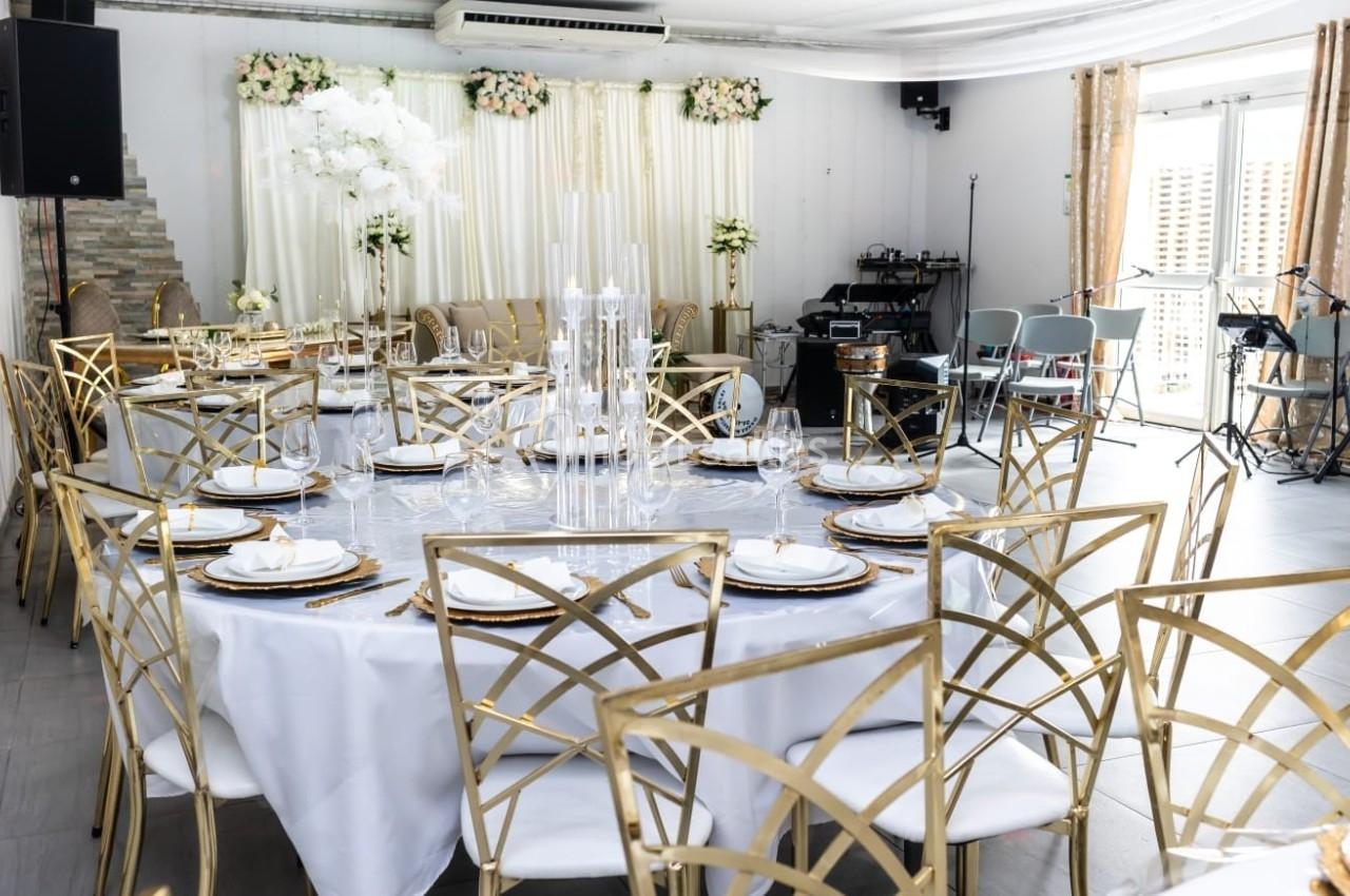 Salle de réception décorée avec des tables rondes, nappes blanches, chaises dorées et arrangements floraux élégants.