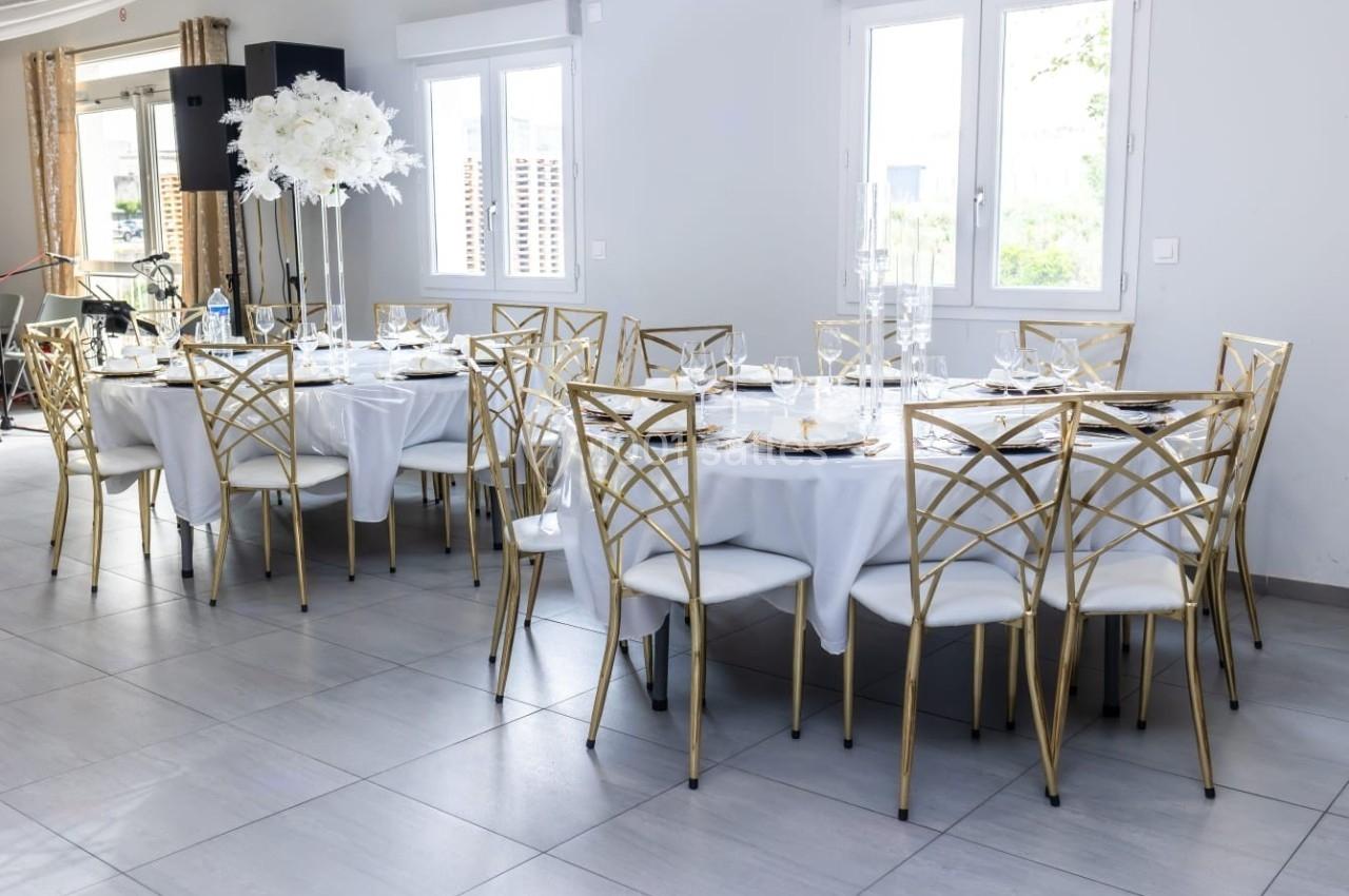 Salle lumineuse avec des tables rondes dressées, nappes blanches, chaises dorées et centre de table floral.