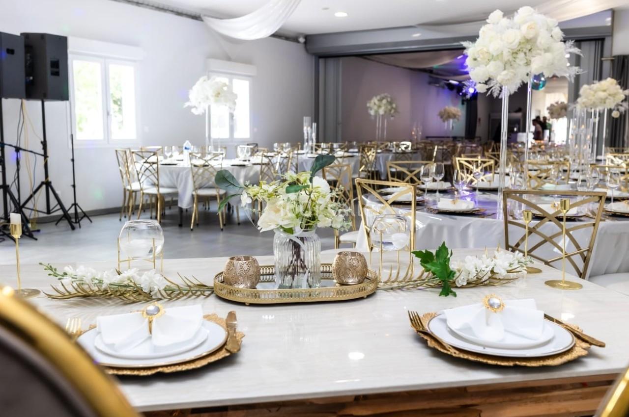 Salle de réception élégante avec tables décorées de fleurs blanches, vaisselle dorée et chaises assorties.