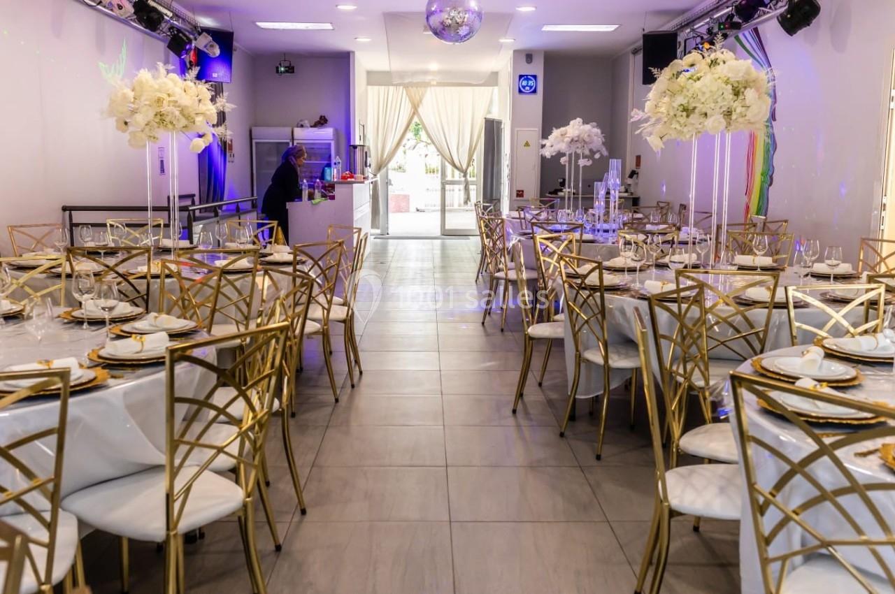 Salle de réception élégante avec tables dressées, chaises dorées et décorations florales blanches.