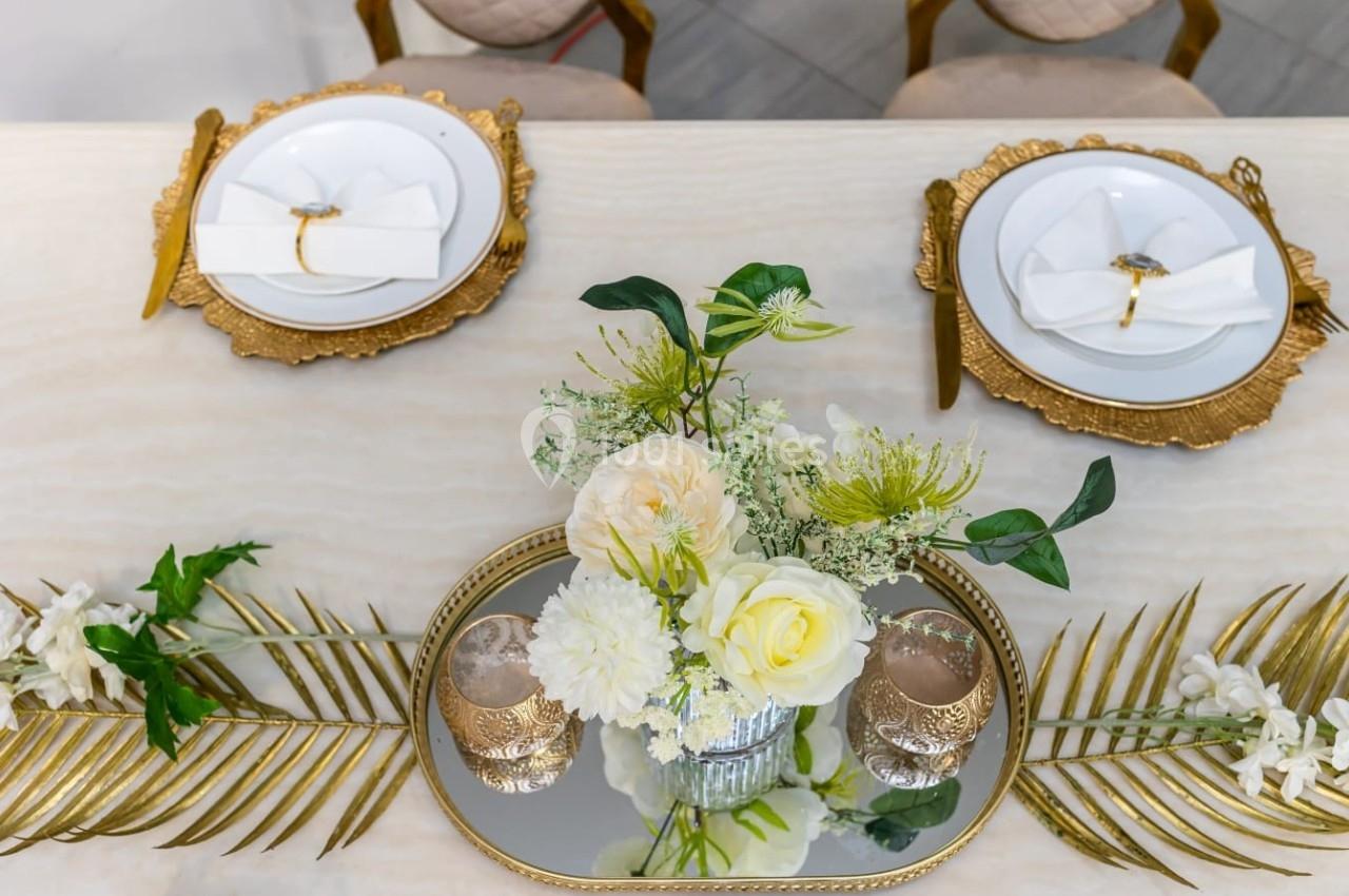 Table décorée avec vaisselle dorée, serviettes pliées, fleurs blanches et jaunes sur un plateau miroir.
